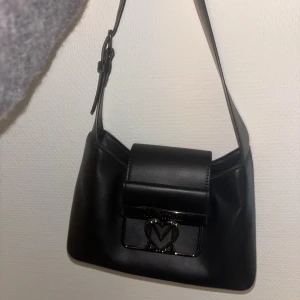 Svart axelväska från Love Moschino - Snygg svart axelväska från Love Moschino i slätt skinnmaterial. Väskan har ett stilrent silverfärgat spänne framtill med logotyp och hjärtformad M-detalj. Justerbar axelrem och enkel, boxig form som ger en modern vibe. Två fack inuti. Nypris runt 2500 kr. 