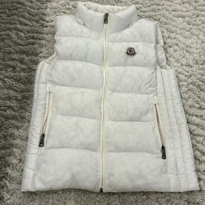 Vit dunväst från Moncler - Säljer en snygg vit dunväst från Moncler med quiltad design och hög krage. Västen har två fickor med dragkedja och Moncler-logga på bröstet. Materialet är polyester och dun, perfekt för kyliga dagar. Stängs med dragkedja framtill.