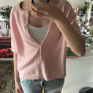 Ljusrosa kofta från 157 med pärlknappar - Superfin ljusrosa kofta från 157 med kort ärm och pärlknappar framtill. Koftan är stickad och har en avslappnad passform, perfekt att slänga över ett linne. Mjuk och mysig känsla, passar till jeans eller kjol.
