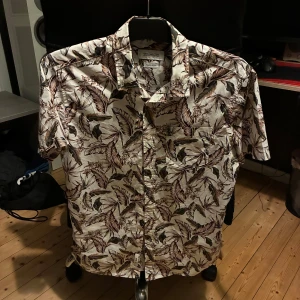 Beige blommig skjorta från Strömberg - Snygg kortärmad skjorta från Strömberg i beige/vit med brunt och svart blommönster. Skjortan är tillverkad i 100% bomull och har klassisk krage samt knäppning framtill. Perfekt för dig som vill sticka ut med ett coolt mönster och skön passform. Använd fåtal gånger 