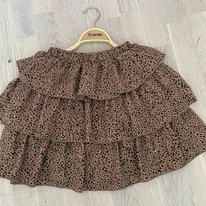 Brun volangkjol med leopardmönster - Supersöt brun kortkjol med flera volanger och svart leopardmönster. Kjolen har resår i midjan för en bekväm passform och är perfekt för dig som gillar trendiga detaljer och djurmönster.