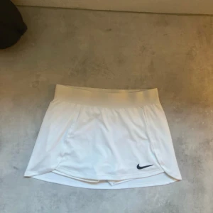 Vit tennis kjol från Nike Dri-Fit - Supersnygg vit kortkjol från Nike med Dri-Fit-teknologi och slim fit-passform. Kjolen har en bred midjeresår och diskret svart Nike-logga framtill. Perfekt för tennis eller träning, tillverkad i ett lätt och svalt syntetmaterial.( Viktigt passar folk som är 137-146cm 