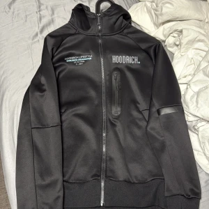 Svart hoodie från Hoodrich XS - Svart hoodie från Hoodrich med tryck på bröstet, ryggen och ena ärmen. Tröjan har hel dragkedja, huva och en bröstficka med dragkedja. Materialet är mjukt och slätt, perfekt för streetwear-vibe.