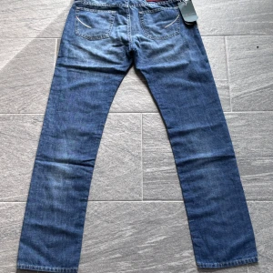 Handpicked Ravello jeans - Nypris 3100 kr. Snygga blå jeans från Handpicked med raka ben och klassisk femficksdesign. Jeansen har röda detaljer vid sömmar och etikett, samt en broderad detalj på bakfickan. Tillverkade i mjukt denimtyg som ger en skön känsla och stilren look.