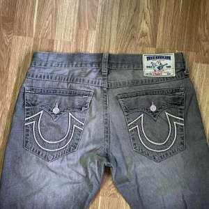 Grå True Religion jeans med kontrastsömmar - Säljer ett par grå jeans från True Religion med raka ben och tydliga vita kontrastsömmar på bakfickorna. Klassisk patch bak i midjan och femficksmodell. Jeansen har en cool faded effekt och är tillverkade i denim.