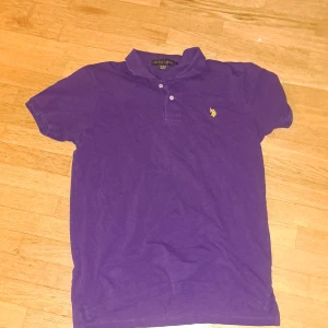 Lila pikétröja från U.S. Polo Assn. - Säljer en lila pikétröja från U.S. Polo Assn. med klassisk krage och två knappar framtill. Tröjan har korta ärmar och en gul broderad logga på bröstet. Perfekt för en chill och stilren look.