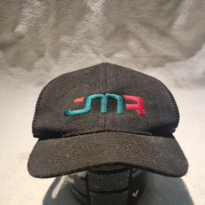 Svart truckerkeps med JMR-brodyr - Svart truckerkeps med böjd skärm och mesh-bakstycke för extra ventilation. Framsidan har broderad JMR-logga i turkos och rött. Justerbar snapback baktill för perfekt passform. Perfekt för en avslappnad streetstyle.