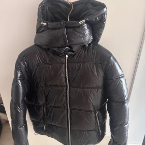 Svart pufferjacka med huva - Säljer en svart pufferjacka med glansig finish och stor huva. Jackan har dragkedja framtill, två fickor med dragkedja och snörning i både huva och nederkant. Perfekt för kalla dagar och riktigt snygg streetstil. Använd 1-2 gånger max! Passar S-M