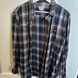 Rutig skjorta från Levi's i blått - Säljer en klassisk rutig skjorta från Levi's i mörkblå, vit och röda toner. Skjortan har långa ärmar, knappar framtill och en bröstficka med Levi's-logga. Perfekt för dig som gillar avslappnad stil med lite edge.