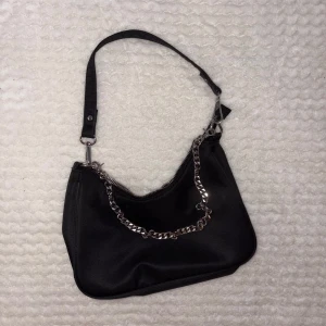 Svart axelväska med kedjedetalj - Stilren svart axelväska med silvrig kedja och dragkedja upptill. Väskan har en enkel, mjuk form och justerbart axelband. Perfekt för dig som gillar minimalistisk och trendig design. Materialet känns slätt och väskan har en cool vibe.
