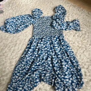 Blå blommig långklänning med puffärm - Supersöt blå långklänning med vita blommor och puffiga långa ärmar. Smockad överdel och voluminösa ärmar ger en trendig look. Perfekt för dig som gillar romantiska vibes och vill sticka ut lite extra.