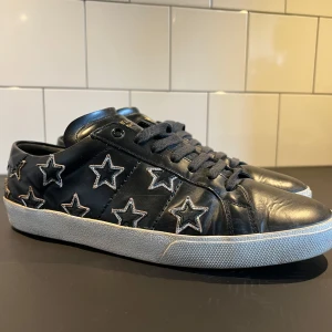 Svarta Saint Laurent sneakers med stjärnor - Svarta sneakers från Saint Laurent med coola broderade stjärnor i vitt längs sidorna. Storlek 43 men passar även 44. Skorna har snörning, vit platt sula och är tillverkade i skinn. Nypris: 6000kr + Mitt pris: 1999