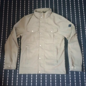 Beige vindjacka från J.Lindeberg S - Stilren beige vindjacka från J.Lindeberg med hög krage, tryckknappar och två bröstfickor med lock. Jackan har raka linjer, diskret logotyp på ärmen och är tillverkad i ett slätt material som känns lätt och smidigt. Perfekt för dig som gillar minimalistisk och clean look. Använd 2 gånger så helt Ny. Ny pris 3400kr