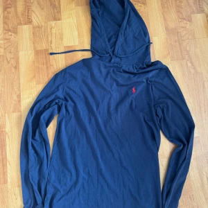 Blå hoodie från Polo Ralph Lauren - Snygg marinblå hoodie från Polo Ralph Lauren med klassisk röd logga på bröstet. Tröjan har huva med snörning och är tillverkad i mjuk bomull. Perfekt för chill dagar och enkel att matcha med jeans eller joggers.