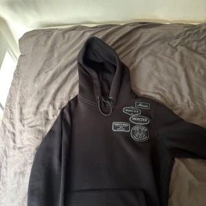 Svart hoodie från Mercier med patches - Svart hoodie från Mercier med flera broderade patches på bröstet. Klassisk huva med snörning och känguruficka framtill. Perfekt för dig som gillar streetwear och vill ha en stilren outfit 