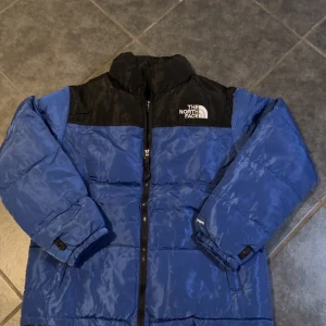 Blå North face pufferjacka - Blå och svart pufferjacka från The North Face med hög krage och dragkedja framtill. Jackan har avtagbara ärmar så den kan bäras som väst. Logga på bröstet och axeln, samt justerbara muddar. Perfekt för kalla dagar och riktigt snygg streetstil.
