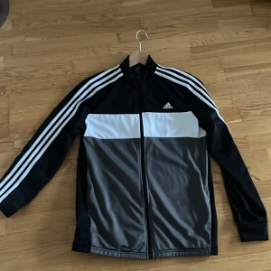 Adidas svart och grå ziptröja - Snygg Adidas ziptröja i svart, grått och vitt med klassiska tre ränder längs ärmarna. Tröjan har hel dragkedja, hög krage och Adidas-logga på bröstet. Perfekt för träning eller chill. Materialet är mjukt och sportigt, troligen polyester. Kom med bud
