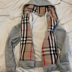 Grå hoodie från Burberry med dragkedja - Grå hoodie från Burberry med klassiskt rutigt foder i beige, svart, vitt och rött. Tröjan har huva med snörning, dragkedja framtill och broderad logga på bröstet. Tillverkad i mjuk bomull med lite elastan för extra komfort.