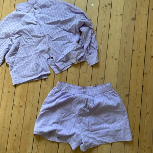 Ljuslila randig skjorta med shorts - Säljer en ljuslila skjorta med matchande shorts. Skjortan har knappar framtill, krage och långa ärmar. Båda plaggen har ett diskret randigt mönster och är tillverkade i ett tunt, skönt bomullstyg. Perfekt för en avslappnad och chill look.