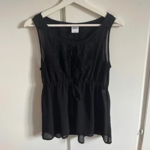 Svart blus med spets från Vero Moda - Svart blus från Vero Moda med spetsdetaljer upptill och knytband i midjan. Blusen är ärmlös och har en lätt, luftig känsla med transparent nederdel. Perfekt för dig som gillar en feminin och stilren look.