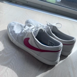 Vita Nike sneakers med rosa swoosh - Säljer ett par vita Nike sneakers med klassisk rosa swoosh på sidan. Skorna har snörning, platt sula och är tillverkade i textilmaterial. Perfekta för dig som gillar en clean och sportig stil med en färgklick. Storlek EU 45.