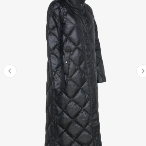 Svart lång quiltad dunjacka - Lång svart dunjacka med quiltat mönster och hög krage. Jackan har raka ärmar, tryckknappar framtill och en oversized passform. Perfekt för kalla dagar när du vill hålla stilen och värmen.