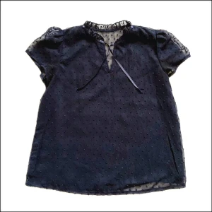 Mörkblå blus med knyt och puffärm - Söt mörkblå blus med korta puffärmar och knytband vid halsen. Blusen har små prickar i tyget och en lätt transparent känsla. Perfekt för dig som gillar romantiska detaljer och en luftig look.