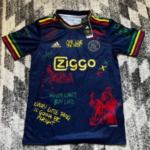 Ajax x Bob Marley Adidas fotbollströja - Ajax x Bob Marley limited edition fotbollströja i mörkblått med färgglada detaljer i rött, gult och grönt. Tryck med Ziggo-logga, Ajax-märke och Bob Marley-grafik samt citat. Korta ärmar med ränder och Aeroready-material för extra komfort.