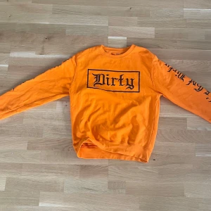 Orange sweatshirt med tryck Dirty - Säljer en orange sweatshirt med svart tryck 'Dirty' på bröstet och text längs ena ärmen. Tröjan har rund halsringning och är tillverkad i mjuk bomull. Perfekt för dig som gillar streetwear och vill sticka ut med en färgstark look.