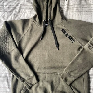 Olivgrön hoodie från MM Sports - Olivgrön hoodie från MM Sports med svart tryck på bröstet. Tröjan har huva med svarta snören, ribbade muddar och detaljerad struktur på ärmarna. Perfekt för chill eller gym. Mjuk insida och klassisk känguruficka framtill.