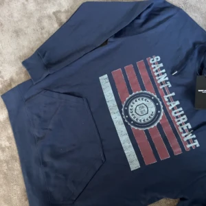 Saint laruent hoodie marinblå (otroligt sällsyn) - Snygg marinblå hoodie från Saint Laurent med tryck i rött och vitt framtill. Tröjan har huva med snörning, magficka och långa ärmar. Perfekt för dig som vill ha en stilren och trendig look med exklusiv känsla.
