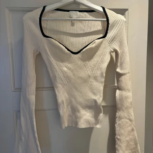 Ribbad vit långärmad topp H&M - Säljer en ribbad, figurnära vit långärmad topp från H&M med svart kant runt den djupa v-ringningen. Tröjan har markerade sömmar framtill och är stretchig och mjuk, med utsvängd arm. 