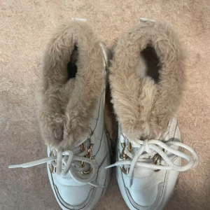 Chunky sneakers med fuskpäls och gulddetaljer - Vita chunky sneakers med grov sula och beige fuskpälsfoder runt öppningen. Skorna har snörning med guldfärgade detaljer och en cool, sportig vibe. Perfekta för dig som vill ha både värme och stil under höst och vinter.