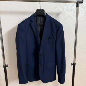 Marinblå kostym från Selected Homme - Snygg marinblå kostym från Selected Homme med klassisk kavaj och matchande kostymbyxor. Kavajen har två knappar, smala slag och fickor med lock. Byxorna har pressveck och bakficka med knapp. Perfekt för dig som vill ha en stilren och modern look. Få två st olika sätt med skjorta med och skor 