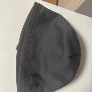 Grå träningsmössa från Under Armour - Mörkgrå mössa från Under Armour med diskret logga framtill. Tillverkad i mjukt syntetmaterial med fleecefoder på insidan för extra värme. Perfekt passform och enkel design, idealisk för träning utomhus under kyliga dagar.