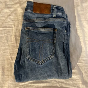 Blåa tiger of sweden jeans - Snygga blå jeans från Tiger of Sweden. Klassisk femficksdesign med bruna detaljer på bakfickan och smal passform. Jeansen är i stretchigt bomullsmaterial och har dragkedjegylf. Perfekta för en stilren och avslappnad look.