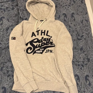 Grå Superdry hoodie med tryck - Grå hoodie från Superdry med stort svart broderat logotyptryck på bröstet och japanska tecken. Tröjan har huva med snörning, ribbade muddar och en klassisk känguruficka. Inga defekter!