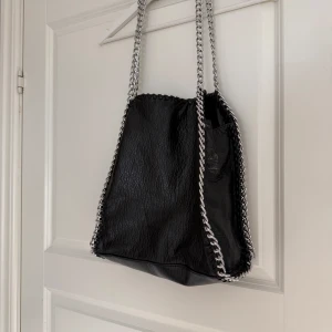 Svart axelväska med kedjedetaljer - Cool svart axelväska i skinnimitation med grova silverfärgade kedjor längs kanterna och som axelrem. Väskan har en mjuk, något slouchy form och en dragkedjeficka på baksidan. Perfekt för dig som gillar edgy detaljer och vill sticka ut.