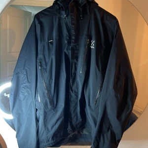 Svart Haglöfs Astral regnjacka M - Svart regnjacka från Haglöfs, modell Astral Jacket Men. Jackan har huva, flera vattentäta dragkedjor och praktiska fickor. Tillverkad i slitstark syntet med minimalistisk design och logga på bröstet. Perfekt för blöta dagar och outdoor.