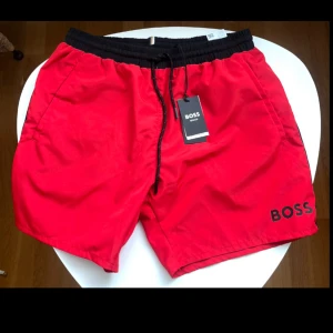Röda badshorts från BOSS med snörning - Snygga röda badshorts från BOSS med svart resårmidja och snörning. Klassisk modell med fickor på sidorna och diskret BOSS-logga framtill. Perfekta för stranden eller poolen, tillverkade i snabbtorkande syntetmaterial. I NYSKICK med prislapp på‼️‼️