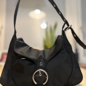 Silfen - Svart axelväska med silverdetalj  - Stilren svart axelväska i syntetmaterial med justerbart axelband och snygg silverfärgad ringdetalj framtill. Väskan har en mjuk, rundad form och lockstängning. Perfekt för dig som gillar minimalistisk och modern design.