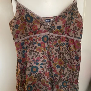 Blommig sommartopp - Söt blommig topp från Active Wear med tunna axelband och knappar framtill. En knapp längst upp har tyvärr lossnat men tyget viker sig inåt vilket resulterar i att man inte märker ”se bild 1). Toppen har en beige bas med färgglada blommor i rosa, blått och gult samt spetsdetaljer längs bysten. Perfekt för varma dagar och enkel att styla.