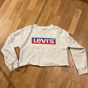 Vit croppad Levi's sweatshirt - Säljer en vit croppad sweatshirt från Levi's med klassisk logga i blått och rött framtill. Tröjan har långa ärmar och rund halsringning. Perfekt för dig som gillar streetstyle och vill ha en enkel men snygg look.