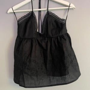 Svart broderad linnetopp från Amisu - Helt nytt svart linnetopp från Amisu med tunna axelband, spetskant vid bysten och broderade detaljer nertill. Toppen har smock i ryggen och knytband i nacken med små silvriga pärlor. Perfekt för en trendig och luftig look.