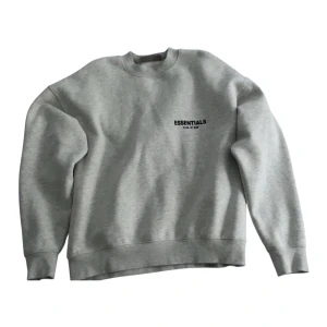 Grå Essentials Fear of God sweatshirt - Säljer en grå sweatshirt från Essentials Fear of God med svart logga på bröstet och ryggen. Tröjan har rund hals, ribbade muddar och är tillverkad i mjuk bomullsblandning. Perfekt för dig som gillar streetwear och enkel stil.
