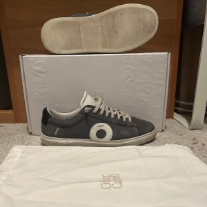 Oliver Cabell low 1 smoke  - Snygga grå sneakers från Oliver Cabell med vit sula och vita detaljer, inklusive en stor vit cirkel på sidan. Skorna har snörning och är tillverkade i skinn med en klassisk låg profil. Perfekta för dig som gillar stilrena och trendiga sneakers.