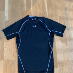 Svart kompressionströja Under Armour - Sportig svart kompressionst-shirt från Under Armour med grå kontrastsömmar och logga på bröstet. Tillverkad i polyester med HeatGear-teknologi som håller dig sval under träning. Kortärmad och tight passform, perfekt för gymmet eller löpning.
