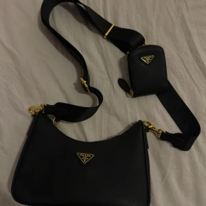 Svart axelväska från Prada med guldiga detaljer - Säljer en svart axelväska från Prada med matchande liten väska på bred axelrem. Väskan har guldiga metalldetaljer, dragkedja och klassisk Prada-logga framtill. Tillverkad i strukturerat skinn och har en modern, stilren form.
