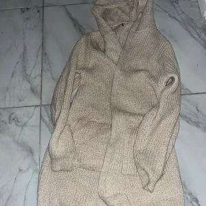 Beige stickad kofta med huva - Mysig beige stickad kofta med huva och stora fickor framtill. Perfekt för lager-på-lager och chill dagar. Koftan har lång ärm och en relaxed passform som gör den superbekväm.