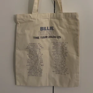 Billie Eilish tygkasse The Tour 2024/25 - Snygg tygkasse i beige med tryck från Billie Eilish The Tour 2024/25. Väskan har blå och svart text med turnéstäder på framsidan. Perfekt för dig som vill visa din kärlek till Billie och ha en praktisk väska till vardagen.
